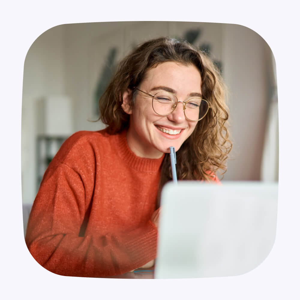 Woman wearing glasses and a red sweater, smiles thoughtfully while holding a pen to her chin and looking at a computer.