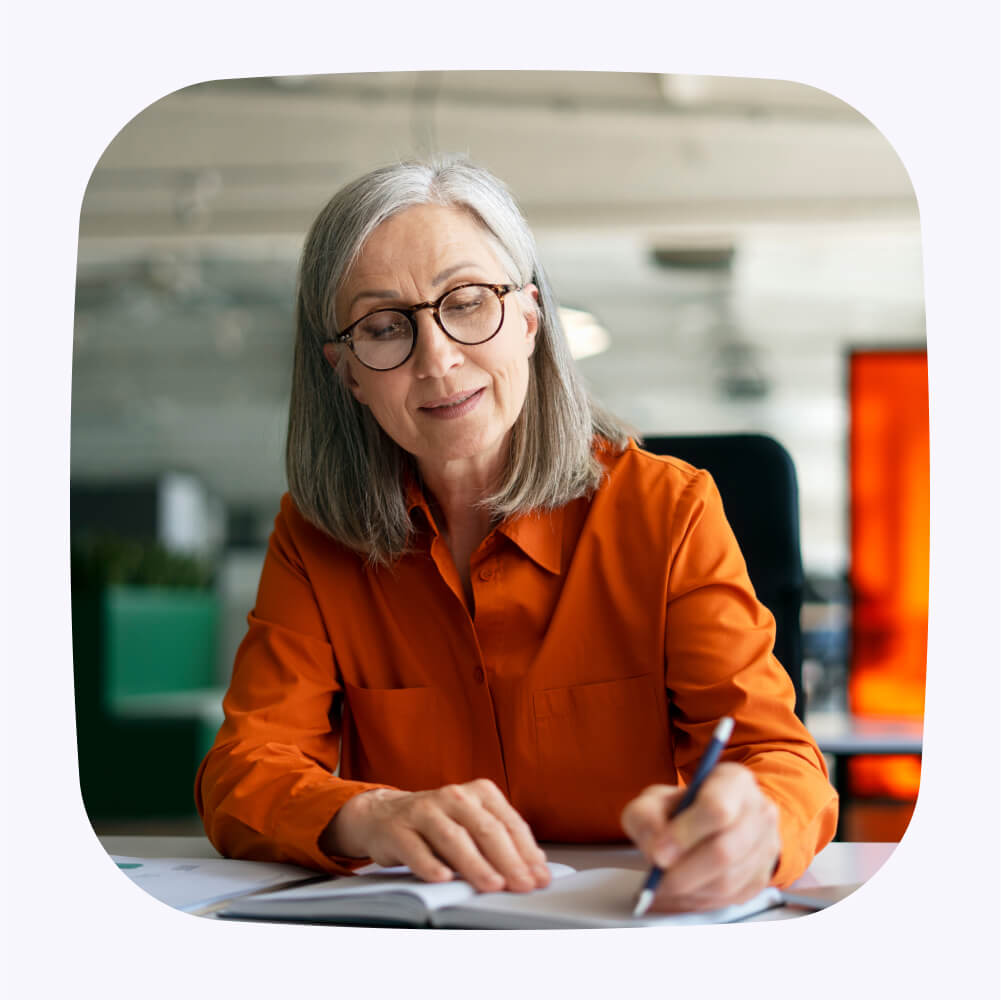 A woman with glasses and an orange collared shirt is writing in a notebook.