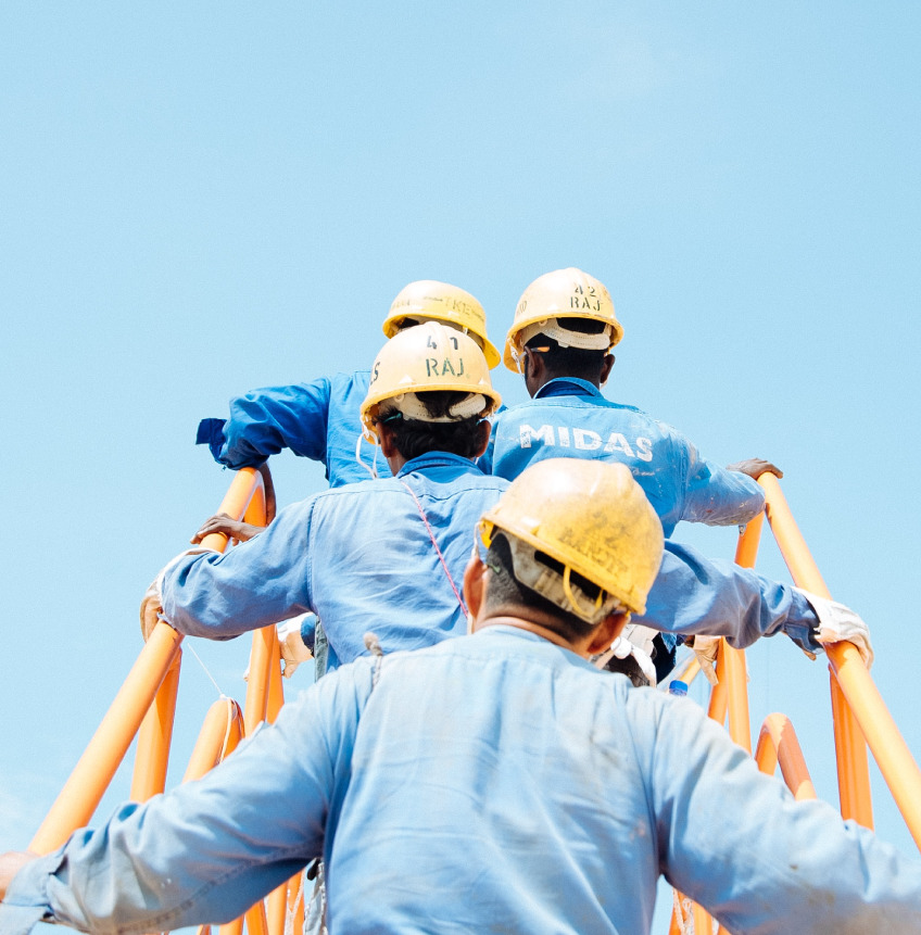 Construction workers in blue uniforms and yellow helmets climbing an orange structure against a clear blue sky