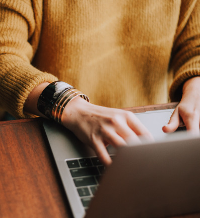 Close-up of a person wearing an orange sweater and bracelet, typing on a laptop.