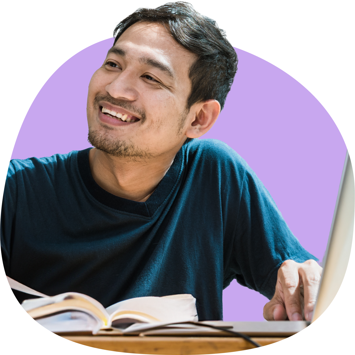 A cheerful man sitting at a desk with an open book and a laptop, smiling and looking up.