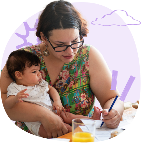 A woman holding a baby while writing on paper, with juice and stationery on the table.