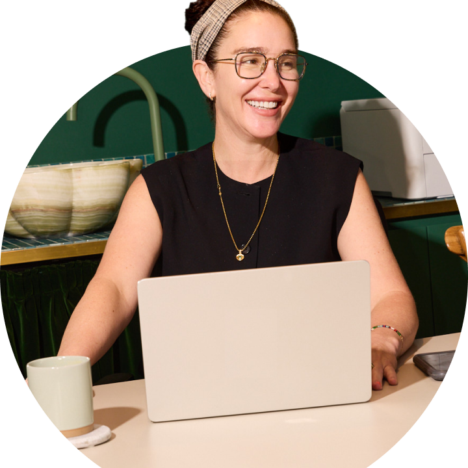 A smiling woman, wearing a black sleeveless top, golden necklace, and eyeglasses, sits in front of her laptop.