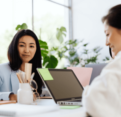 Two professional women are in a bright office, working on laptops and discussing something. The space is modern, with plants and office supplies around.