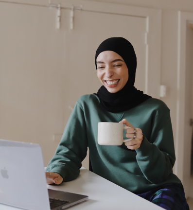 Smiling woman wearing a hijab and green top, holding a coffee mug while facing her laptop.