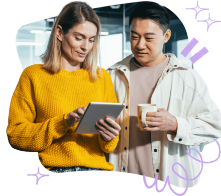 A woman in a yellow sweater holds a tablet as a man in a beige jacket, holding a coffee mug, looks at the screen.