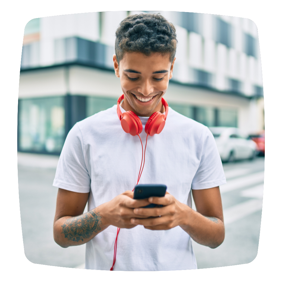 A young man with short curly hair, wearing a white t-shirt and red headphones around his neck, smiling while looking at his phone